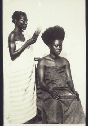 Two Black women. One is sitting. The other is standing while picking the hair of the one sitting